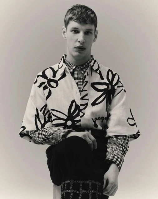 Monochromatic portrait of a young adult male model wearing a floral print shirt, plaid shirt, and dark pants in a light studio setting.
