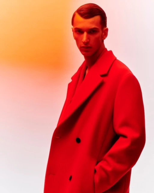 Adult male poses in a red overcoat with an orange and white gradient backdrop in a fashion-forward portrait-style photograph.
