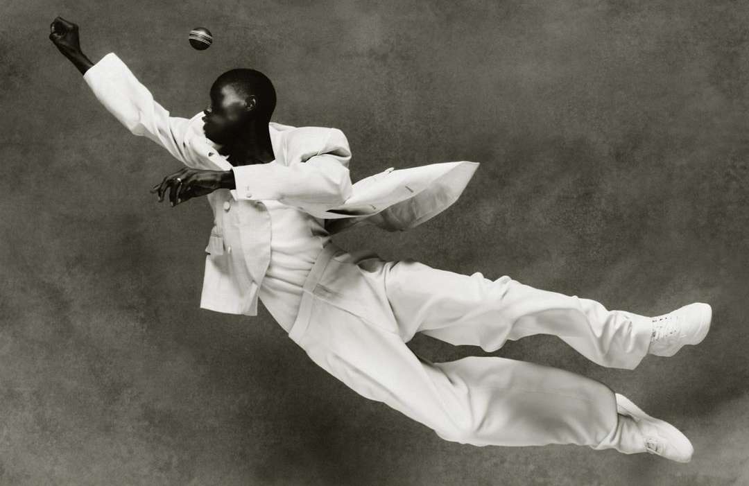 A person in a white suit is mid-air reaching for a ball, captured in a monochrome photograph with a textured background, fashion forward and dynamic.