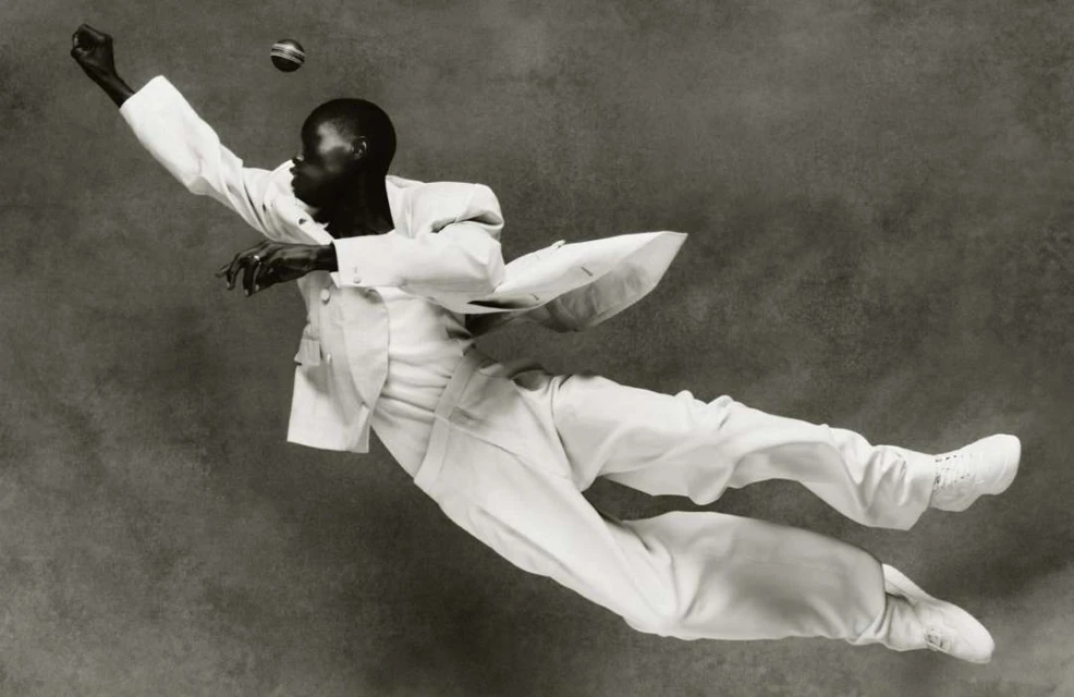 A person in a white suit is mid-air reaching for a ball, captured in a monochrome photograph with a textured background, fashion forward and dynamic.