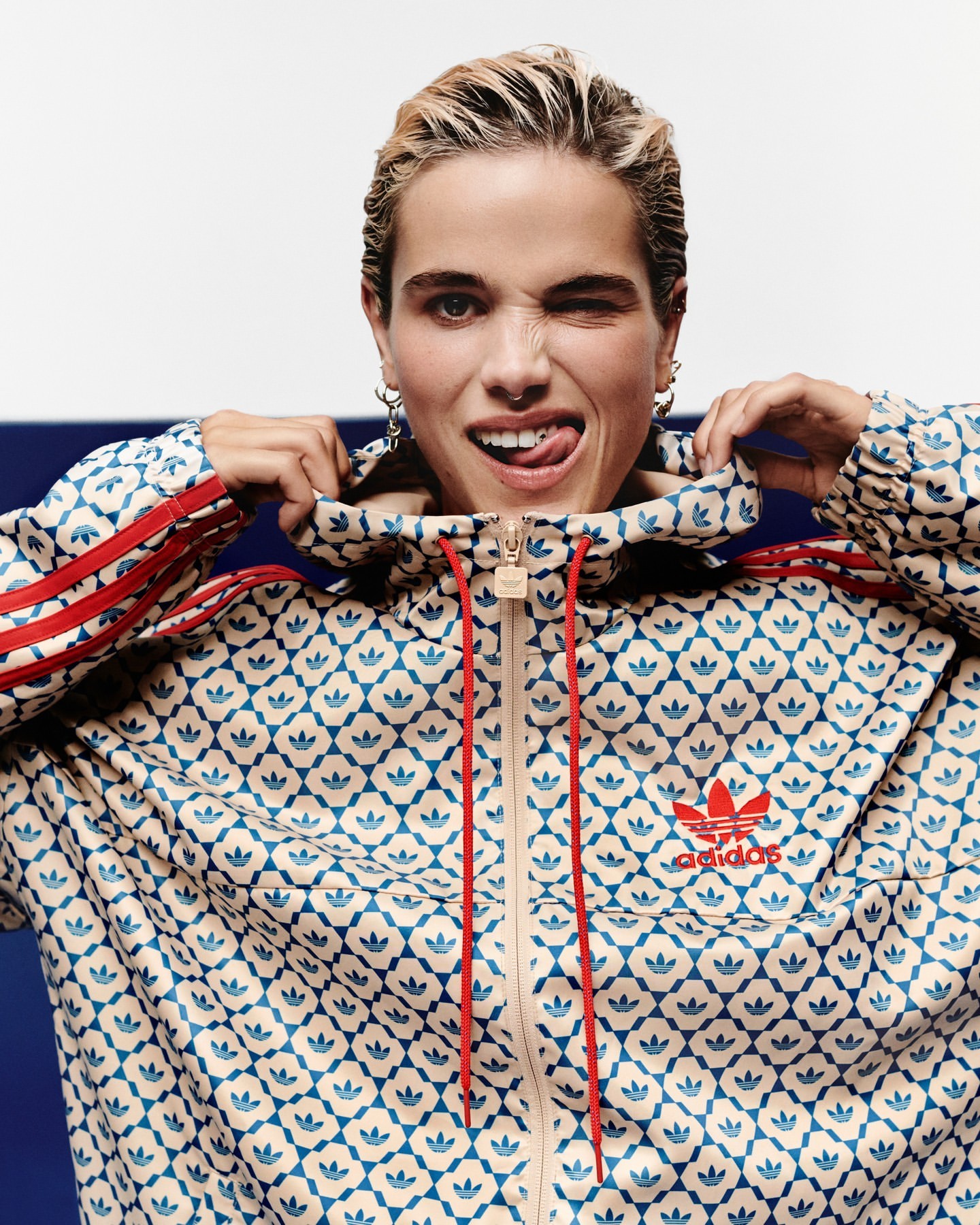 Woman with blonde hair in patterned Adidas tracksuit winking at the camera, showing her tongue and wearing silver hoop earrings.