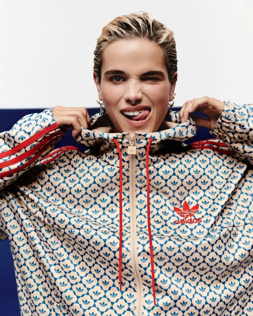 Woman with blonde hair in patterned Adidas tracksuit winking at the camera, showing her tongue and wearing silver hoop earrings.