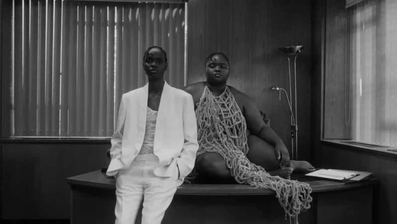 Two Black women in a monochrome office setting. One woman wears a white pantsuit, the other is draped in a woven textile.