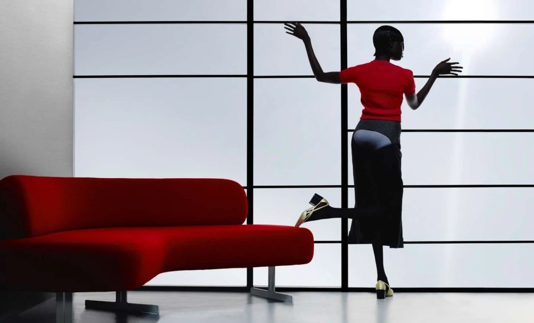 A woman in red strikes a pose beside a red couch in front of a minimalist white and black window, creating a striking visual contrast.