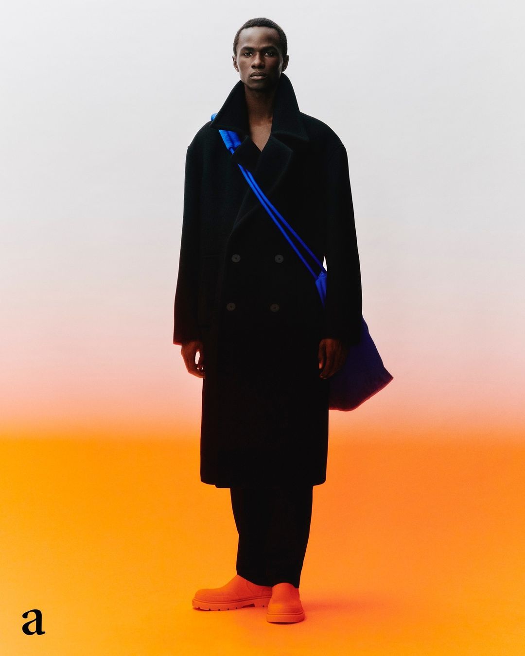 Male fashion model posing in a black double-breasted overcoat with a blue shoulder bag against an orange gradient backdrop.