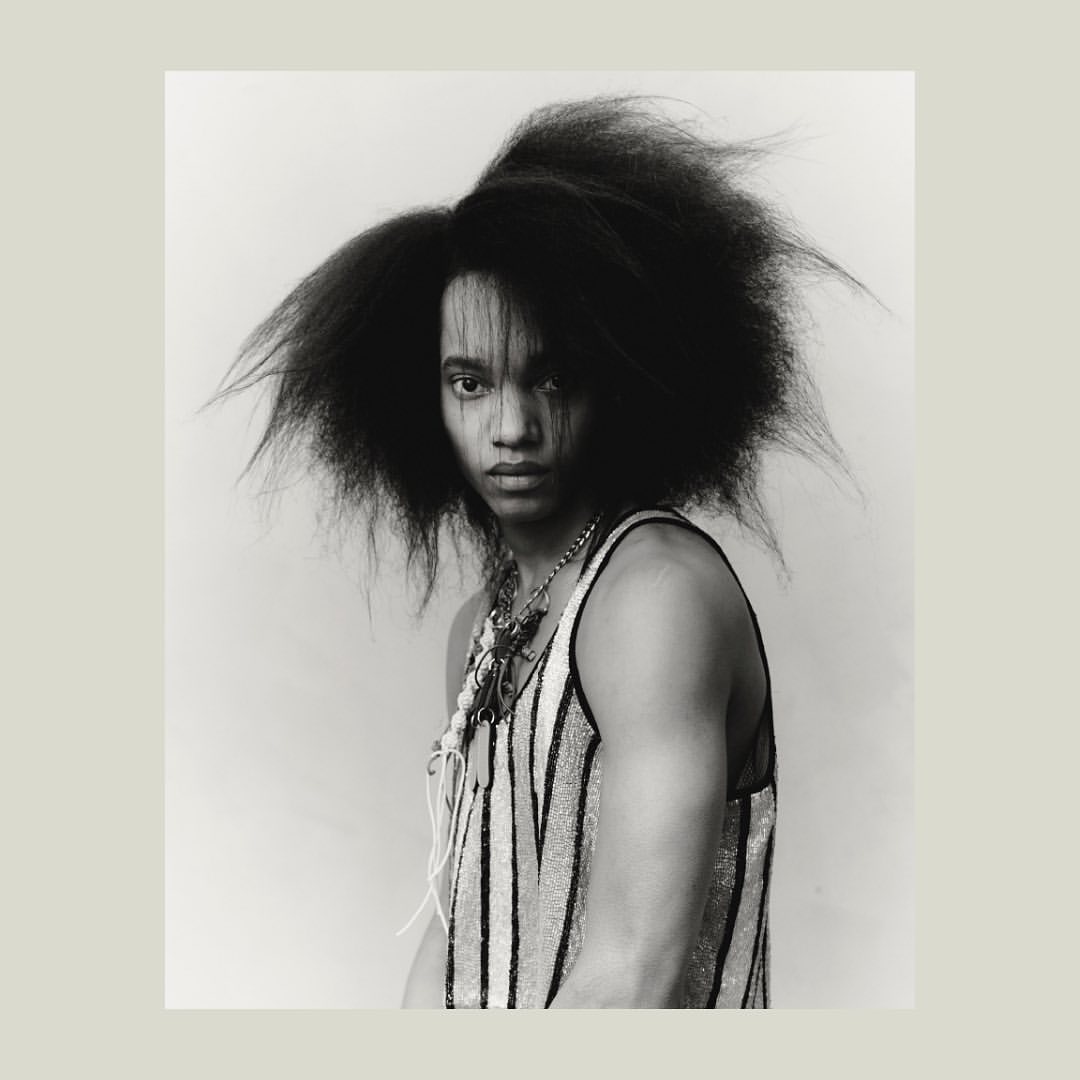 Striking black and white portrait of a young adult with dark hair, wearing a striped tank top and multiple necklaces, against a light backdrop.