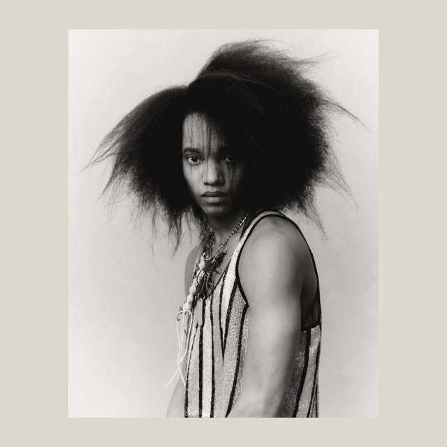 Striking black and white portrait of a young adult with dark hair, wearing a striped tank top and multiple necklaces, against a light backdrop.