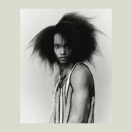 Striking black and white portrait of a young adult with dark hair, wearing a striped tank top and multiple necklaces, against a light backdrop.