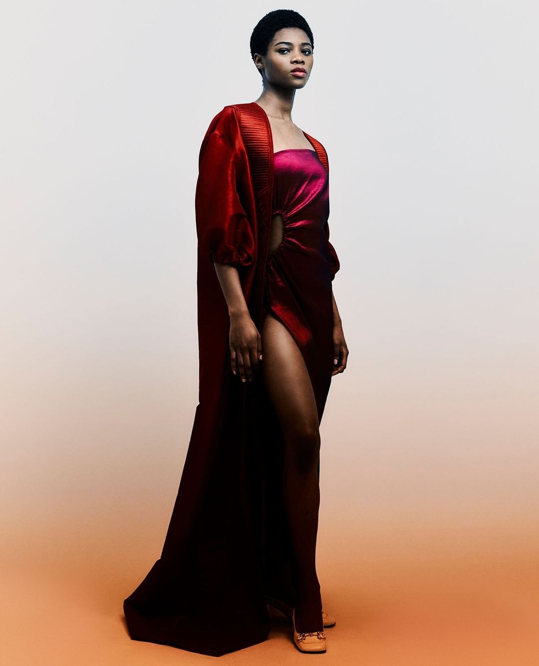 Model in long red coat and magenta dress with high slit against a warm gradient background