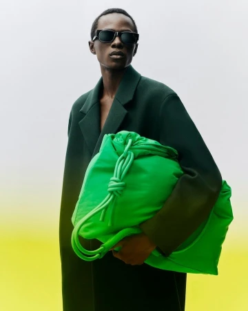 Fashionable person in sunglasses and a green coat holds a large bright green drawstring bag against a white and yellow background.