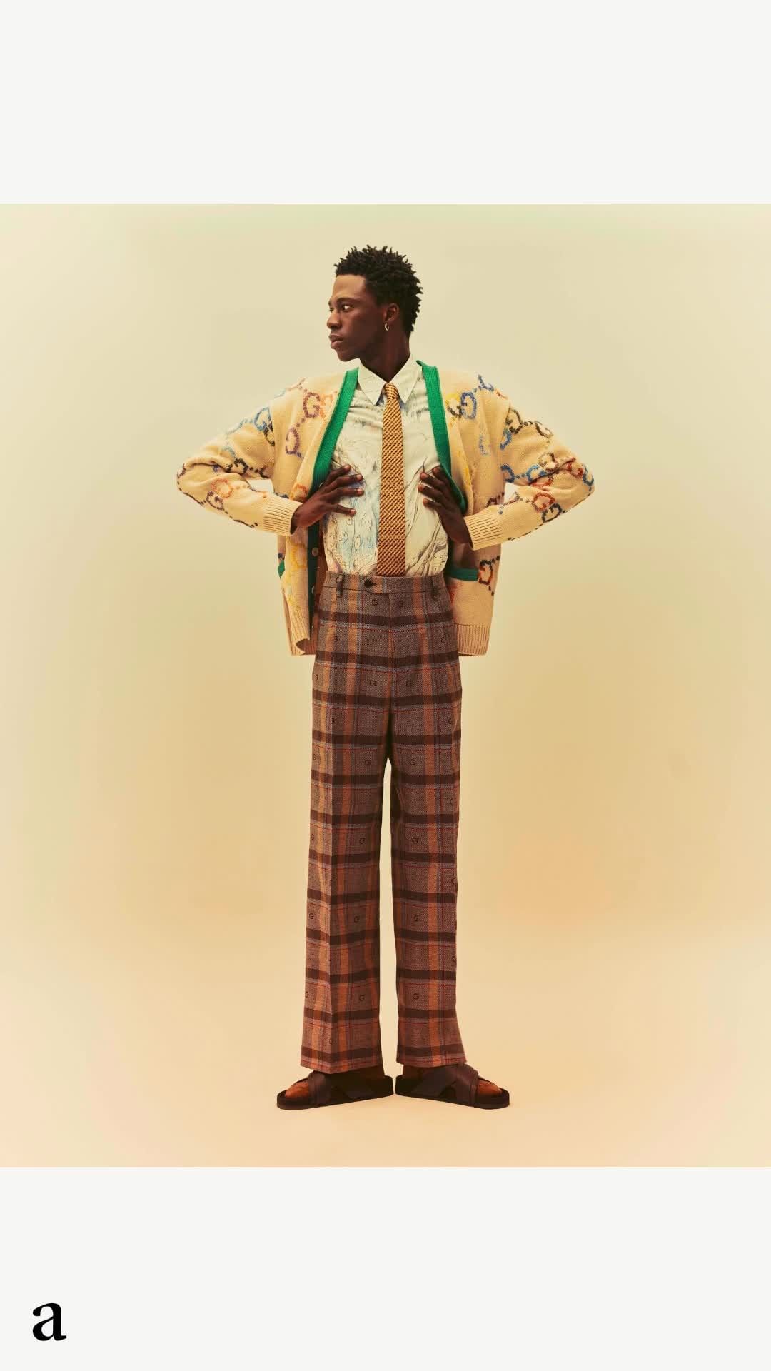 Full-length shot of a Black male model posing in a Gucci cardigan, patterned shirt and trousers in a cream-colored studio setting.