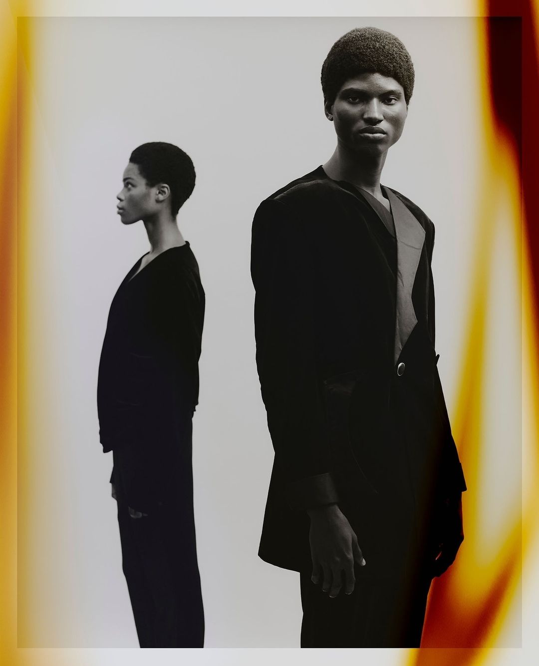 Black man and black woman in dark clothing posing against a light background with warm film burn borders.
