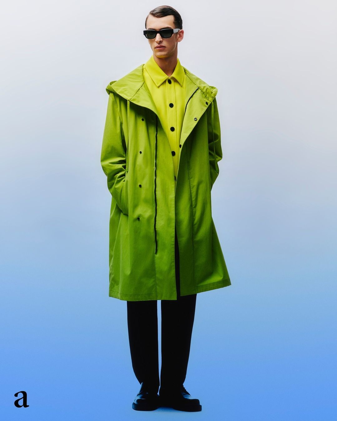 Full shot of a man wearing a green hooded raincoat, sunglasses, yellow shirt, and black pants against a gradient background.
