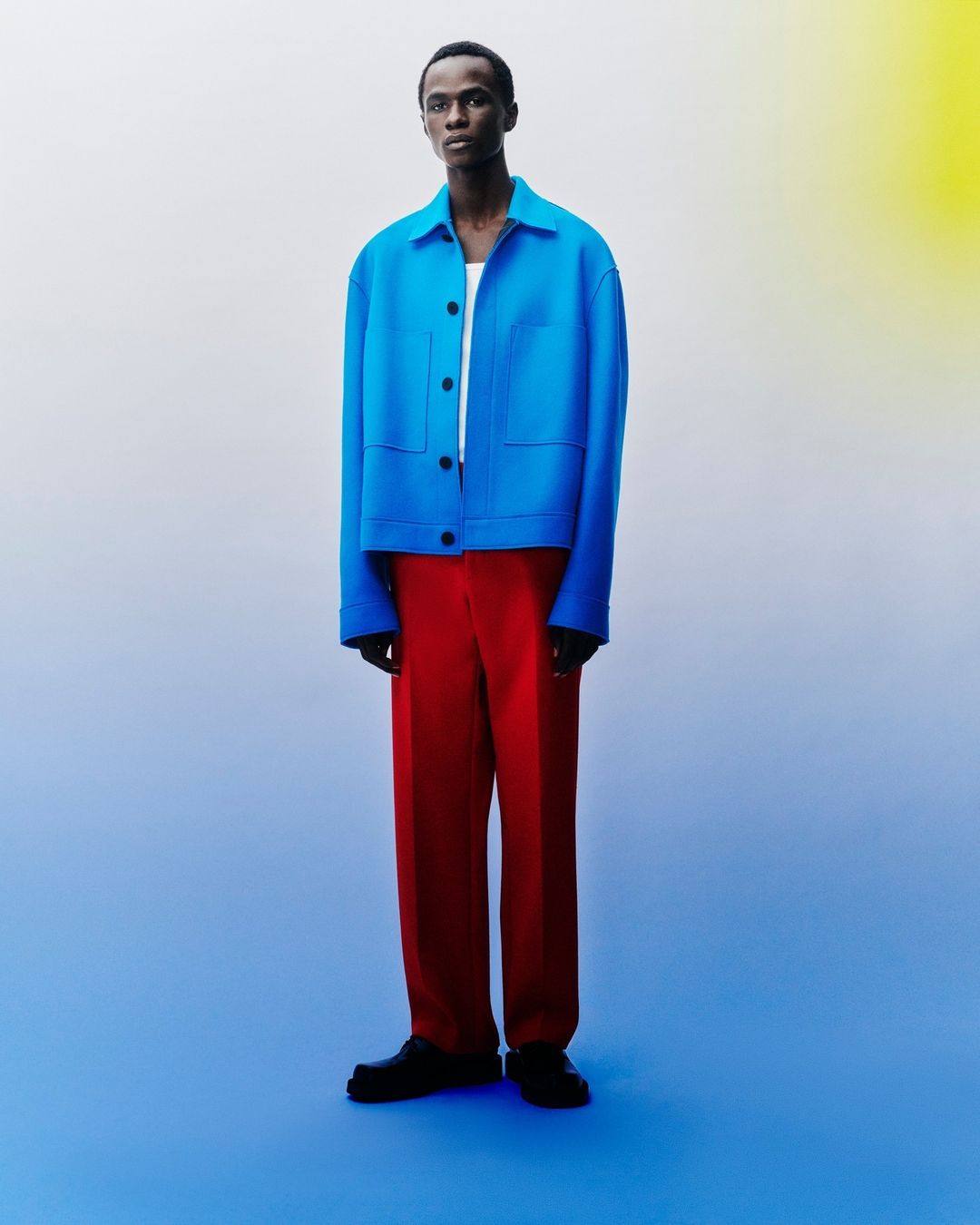 Full-length shot of a male model in a blue jacket and red trousers standing in a blue and white gradient studio background.