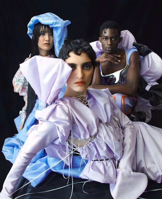 Three diverse fashion models wearing extravagant lilac and blue ruffled outfits pose indoors against a dark background in a studio shot.