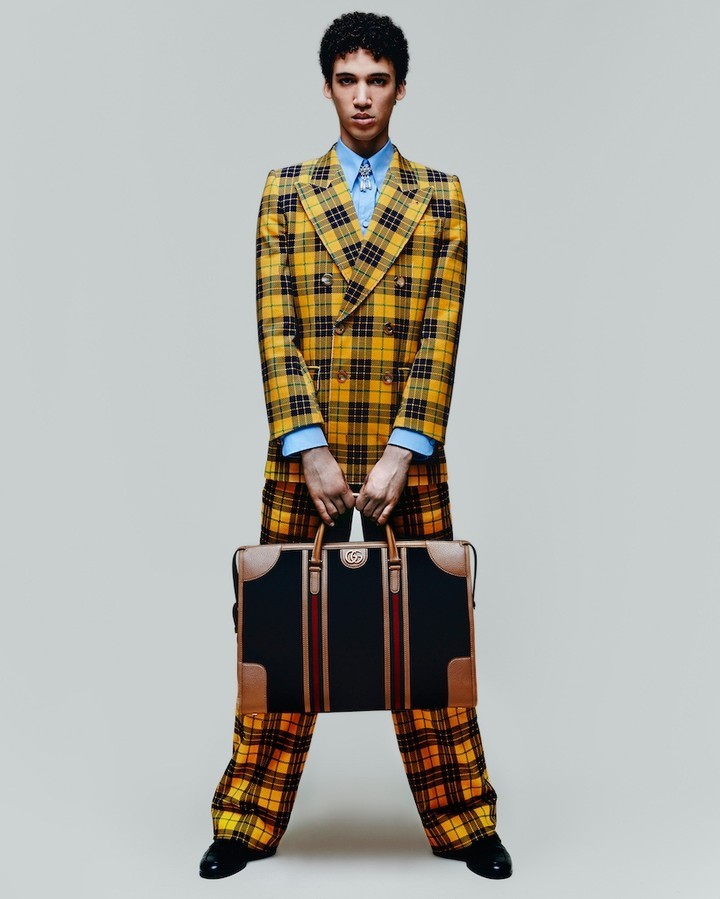 A young adult male model poses in a yellow plaid suit holding a leather briefcase in a studio shot with a neutral background.