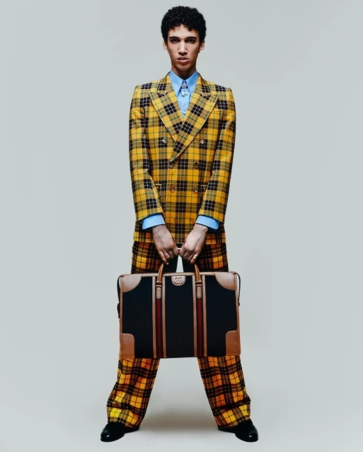 A young adult male model poses in a yellow plaid suit holding a leather briefcase in a studio shot with a neutral background.