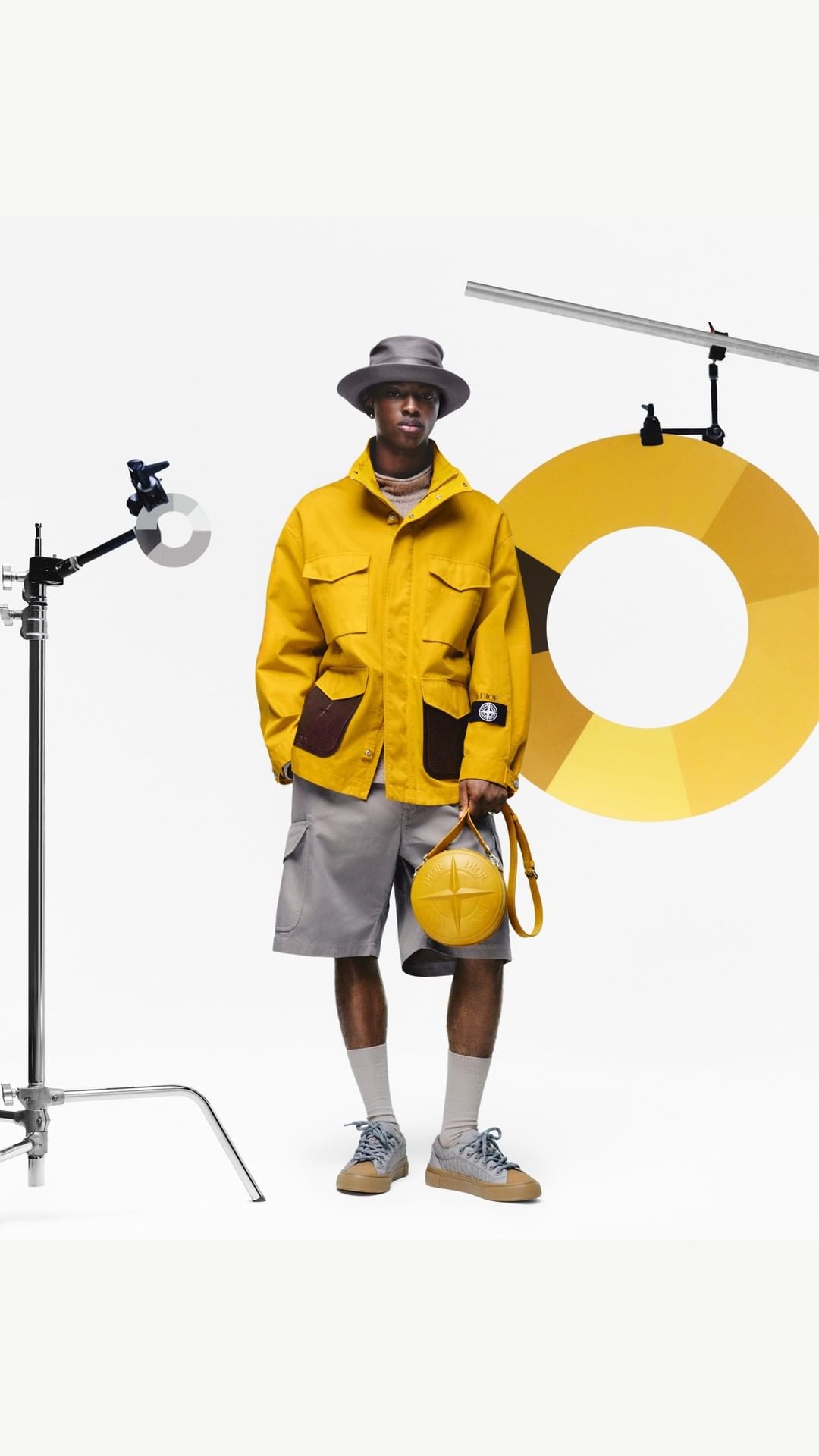 A dark-skinned male fashion model in a yellow jacket, grey shorts, and hat holds a yellow ball-shaped purse against a white backdrop.