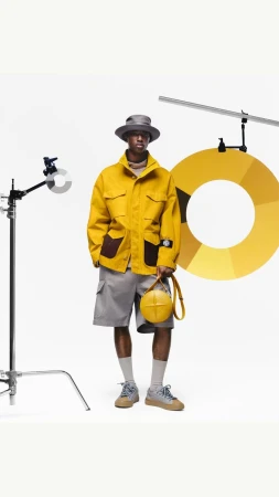 A dark-skinned male fashion model in a yellow jacket, grey shorts, and hat holds a yellow ball-shaped purse against a white backdrop.