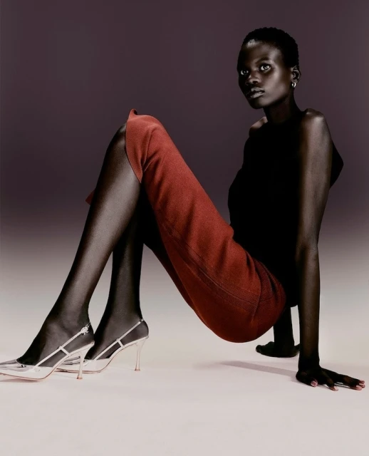 Elegant studio fashion shot of a slender Black woman in rust pants and high heels, gazing at the camera with soft lighting and a neutral backdrop.