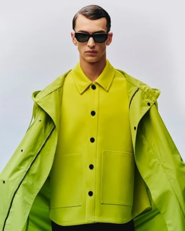 Man wearing black sunglasses and a bright lime green buttoned jacket against a pale grey seamless background.