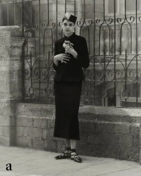 Model in black jacket and skirt holding a flower standing before a decorative iron fence in London by Ethan Hart.
