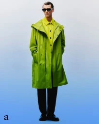 Man wearing a lime green hooded coat over a yellow shirt and dark sunglasses against a blue gradient background.