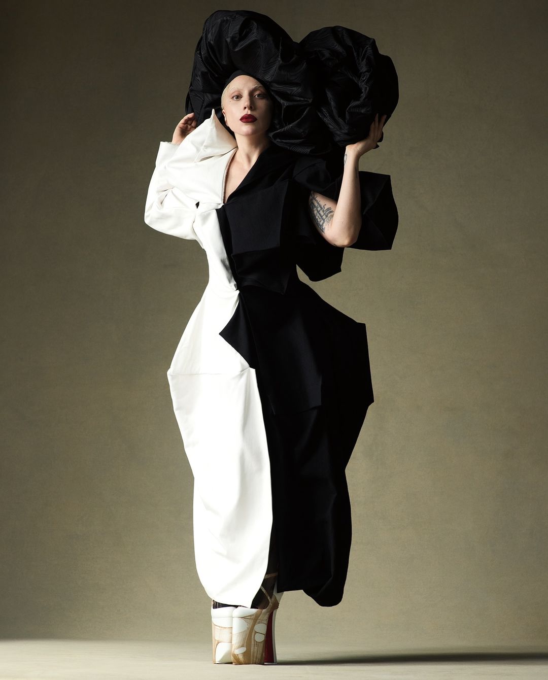 Woman poses in a dramatic black and white dress with a large black headpiece against a neutral backdrop in a fashion forward editorial photograph.