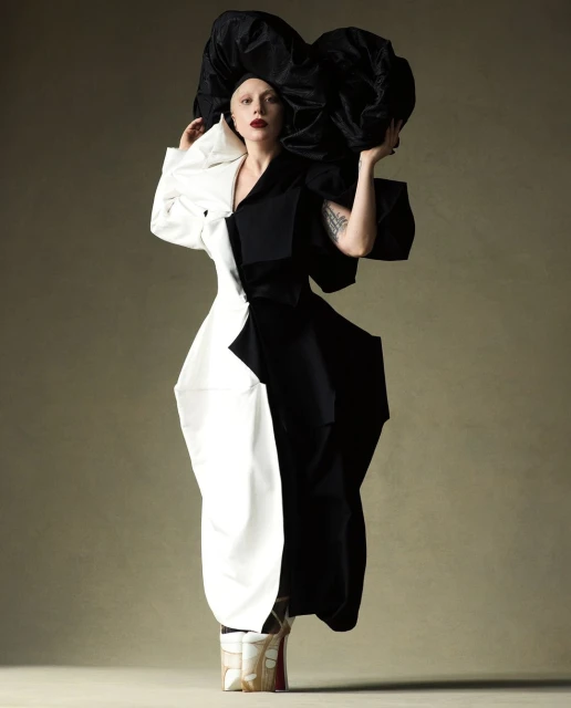 Woman poses in a dramatic black and white dress with a large black headpiece against a neutral backdrop in a fashion forward editorial photograph.