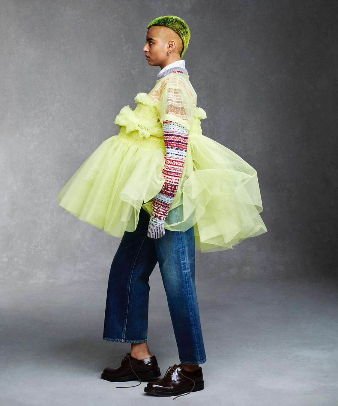 Profile view of a person with a green mohawk, wearing a ruffled yellow dress, striped sleeves, denim pants, and brown dress shoes.