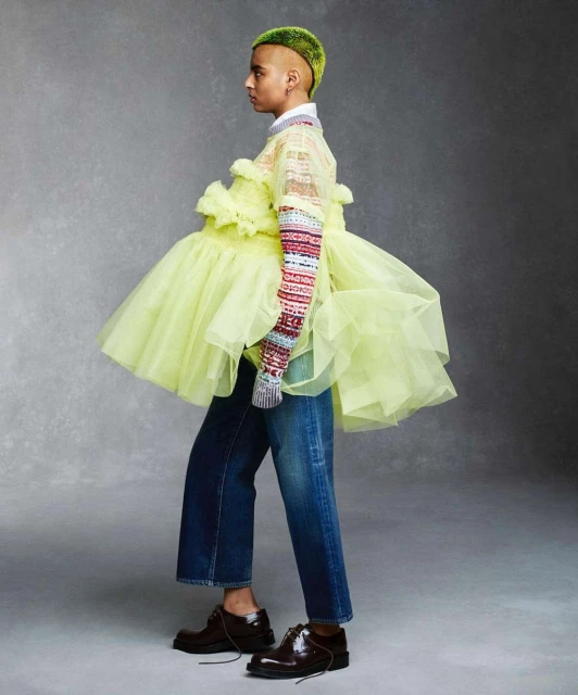 Profile view of a person with a green mohawk, wearing a ruffled yellow dress, striped sleeves, denim pants, and brown dress shoes.