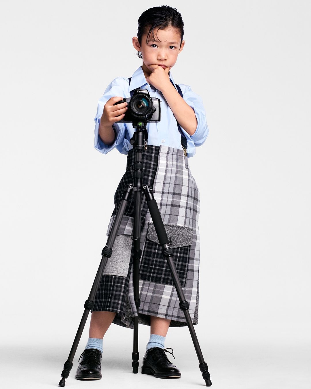 Young child in a light blue shirt and plaid skirt standing behind a professional camera on a tripod