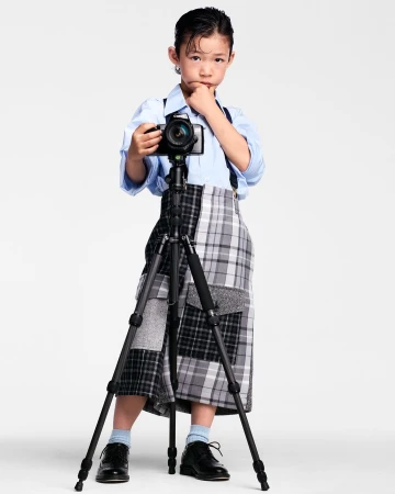 Young child in a light blue shirt and plaid skirt standing behind a professional camera on a tripod