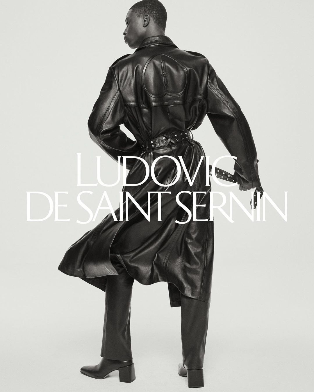 Editorial fashion shot of a Black male model in a black leather coat and pants. He has his back to the viewer in a studio environment.