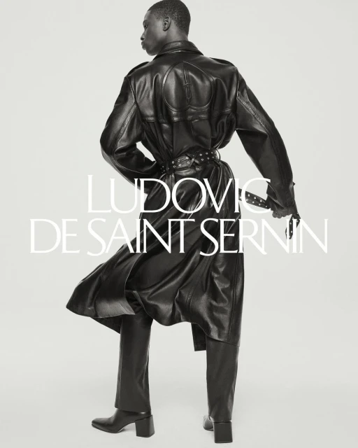 Editorial fashion shot of a Black male model in a black leather coat and pants. He has his back to the viewer in a studio environment.