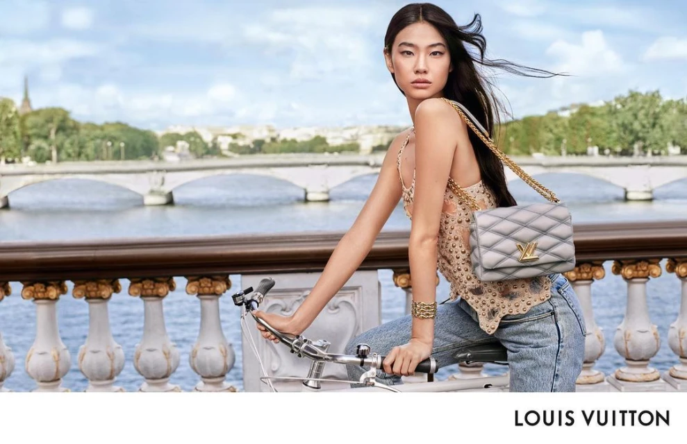 Asian woman wearing designer top and jeans poses with a Louis Vuitton bag while riding a bicycle near a river in an outdoor shot.