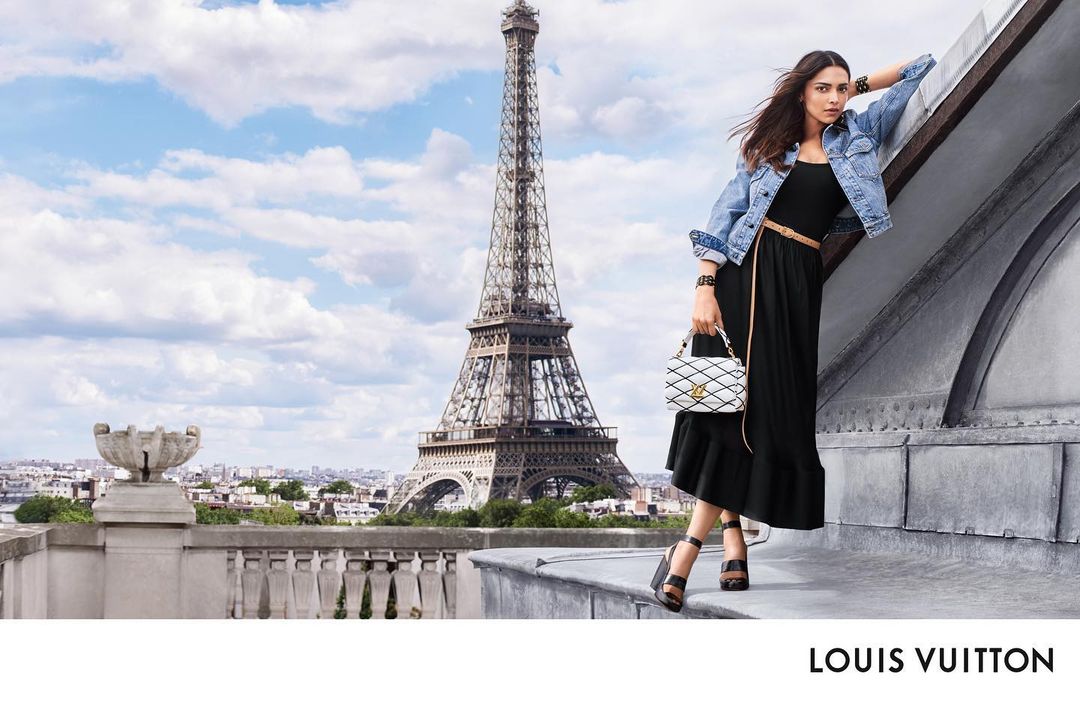 Woman in black dress and denim jacket leaning on a rooftop ledge with the Eiffel Tower in the background