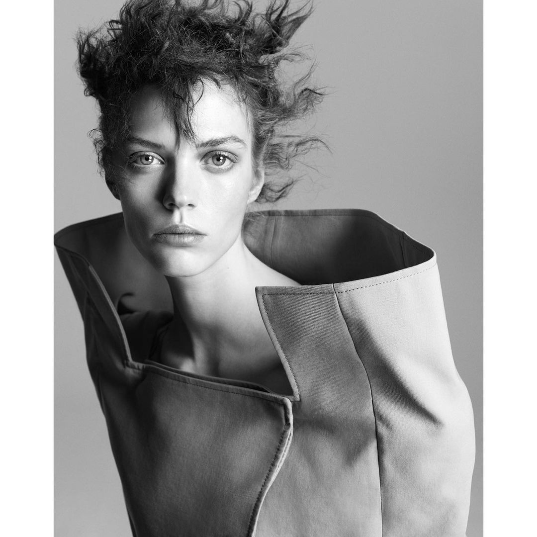 Monochrome portrait of a woman with tousled hair wearing an oversized jacket with a wide collar, gazing directly at the camera.