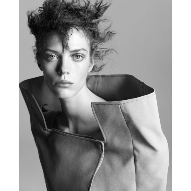 Monochrome portrait of a woman with tousled hair wearing an oversized jacket with a wide collar, gazing directly at the camera.