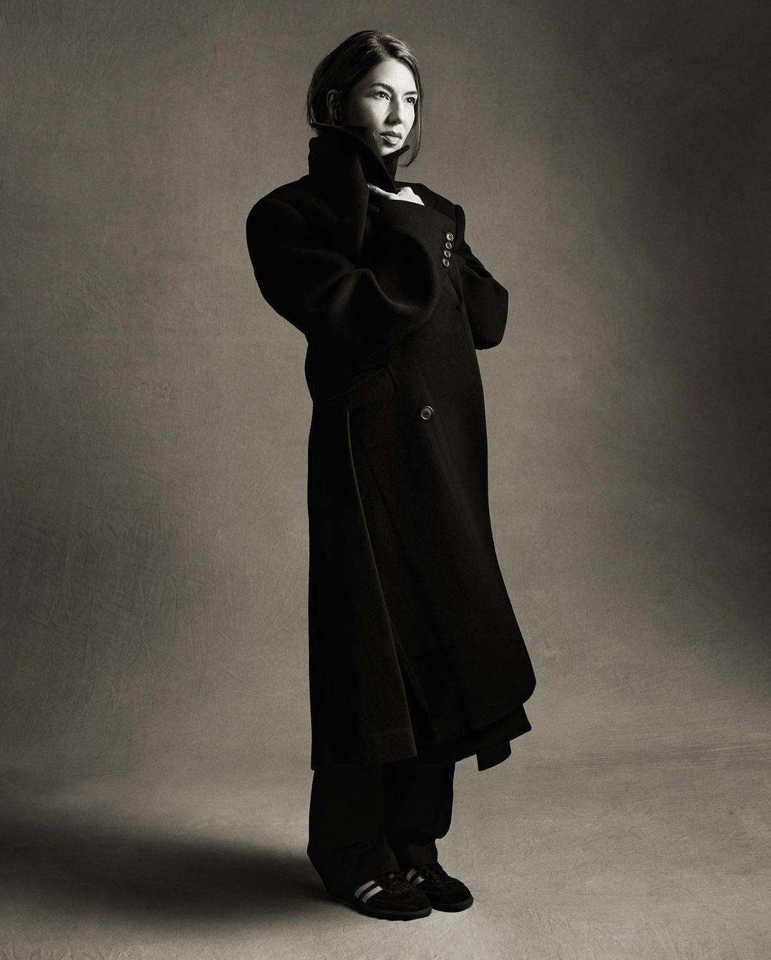 A woman in a long black coat stands in a studio with gray background. Her hands are near her face. The image is in black and white.