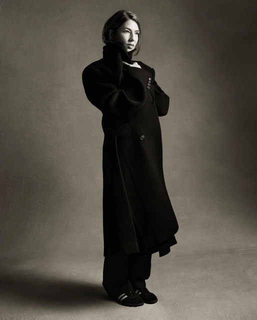 A woman in a long black coat stands in a studio with gray background. Her hands are near her face. The image is in black and white.