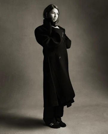 A woman in a long black coat stands in a studio with gray background. Her hands are near her face. The image is in black and white.