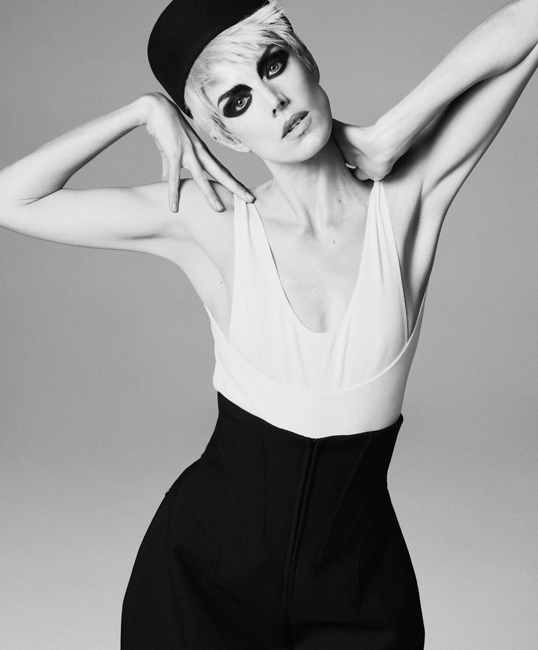 Fashion portrait of an adult woman with short blonde hair and dark eye makeup wearing a white tank top and black pants against gray background.