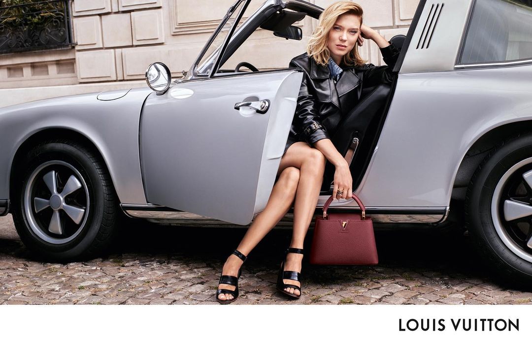Fashion ad featuring a blonde woman in a black leather jacket posing in a silver convertible with a red Louis Vuitton handbag outdoors.