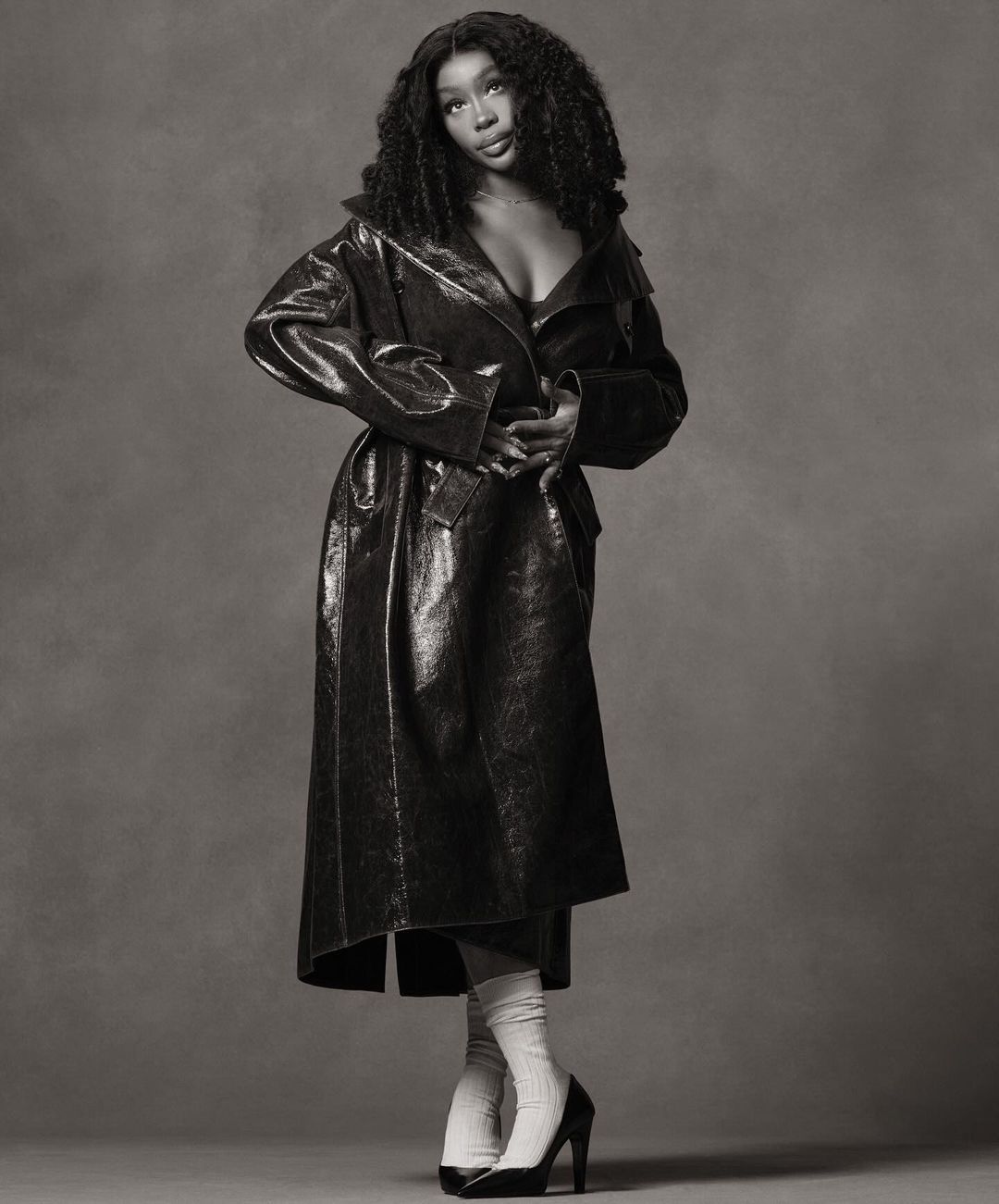 Black and white studio portrait of a Black female model wearing a long vinyl trench coat, tube socks, and high heels against a grey backdrop.