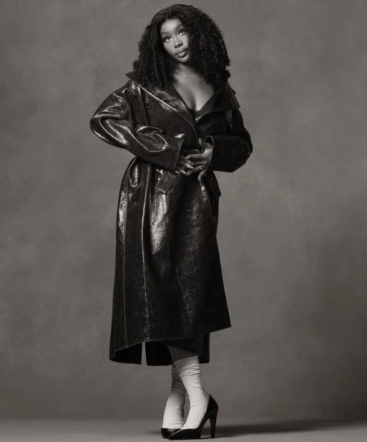 Black and white studio portrait of a Black female model wearing a long vinyl trench coat, tube socks, and high heels against a grey backdrop.