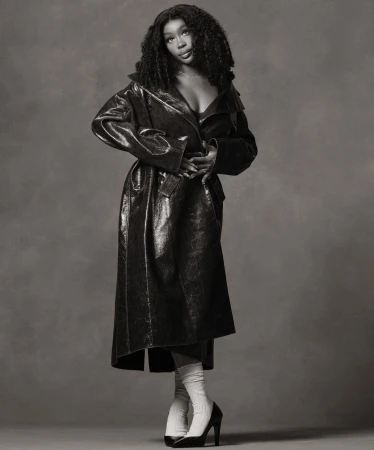 Black and white studio portrait of a Black female model wearing a long vinyl trench coat, tube socks, and high heels against a grey backdrop.