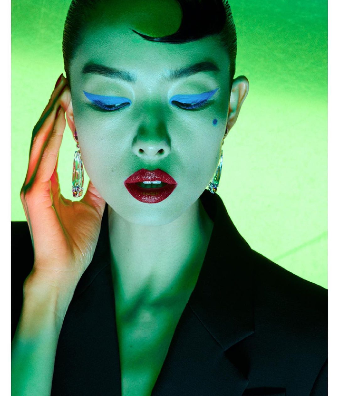 Close-up of a stylish adult woman with graphic blue eye makeup, dark red lipstick, statement earrings, and a black blazer under green lighting.