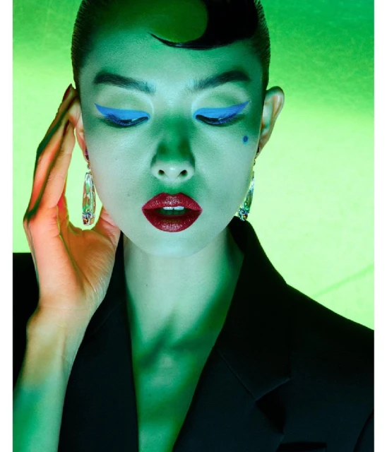 Close-up of a stylish adult woman with graphic blue eye makeup, dark red lipstick, statement earrings, and a black blazer under green lighting.