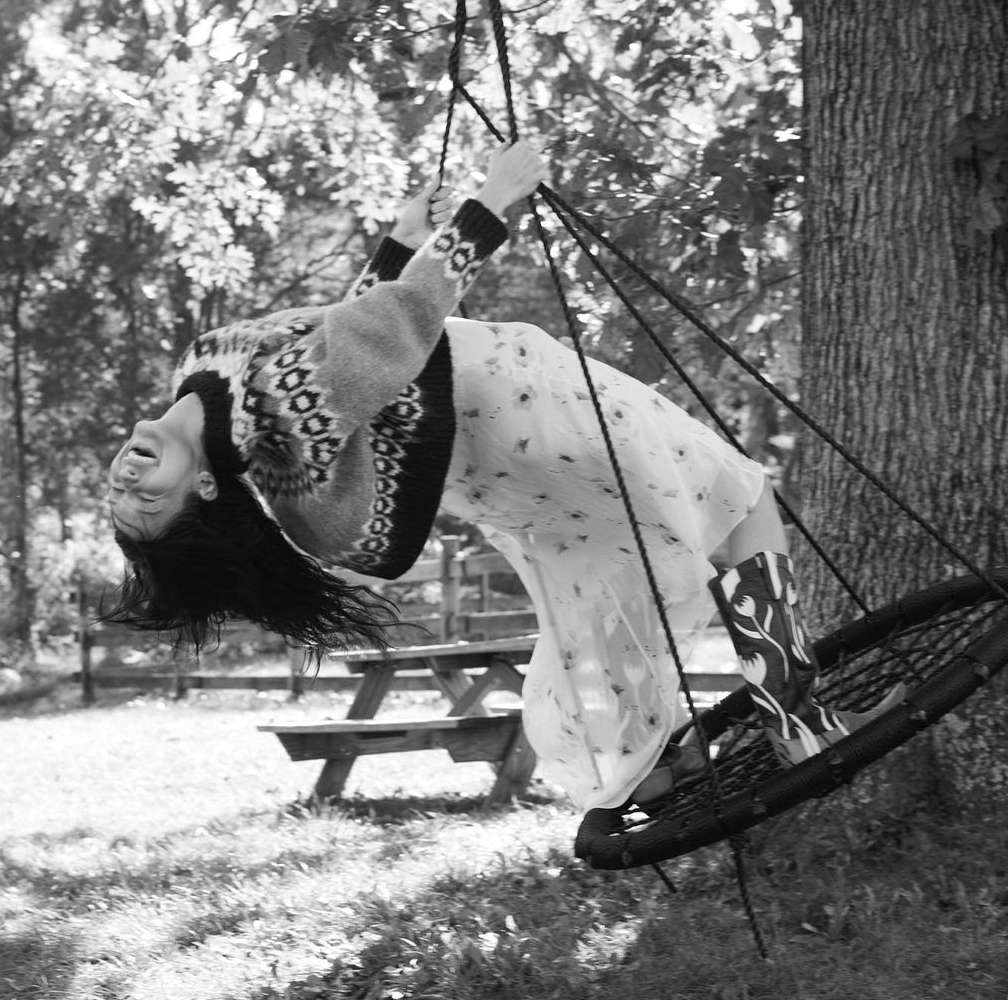 Monochrome image of a woman on a swing, laughing with her head tilted back, in a dress, sweater, and boots in an outdoor setting.